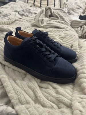 Mörkblå sneakers från Christian Louboutin - Snygga mörkblå sneakers från Christian Louboutin i mocka med klassisk röd sula. Skorna har rund tå, platt sula och snörning. Insidan är fodrad i beige läder och baksidan har broderad logga. Perfekta för dig som vill sticka ut med exklusiva detaljer.