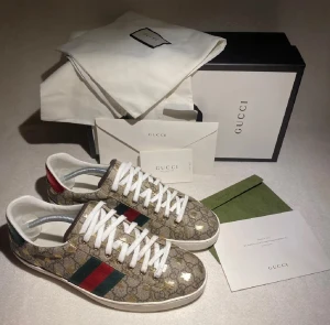 Gucci sneakers med GG-mönster - Säljer ett par lyxiga Gucci sneakers med klassiskt GG-monogram i beige och brunt, dekorerade med guldiga detaljer. Skorna har vita snören, platt sula och rund tå. Sidorna pryds av gröna och röda ränder, och hälen har metallicröd och metallicgrön finish med Gucci-logga. Perfekt för dig som gillar statement pieces.