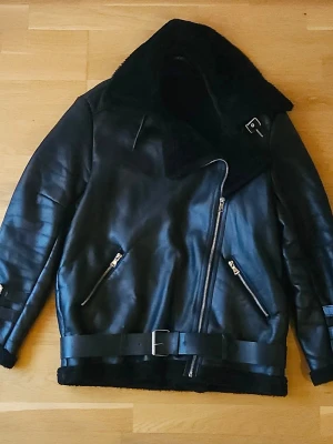 Svart fodrad  jacka - Svart jacka i skinnimitation med bred krage i fuskpäls. Fodrad för extra värme och står emot regn, perfekt för kyligare höstdagar eller varmare vinterdagar. Fickorna har släppt lite i sömmarna men är lagade.