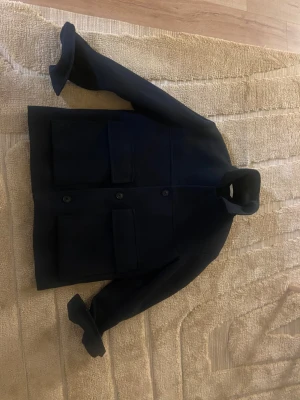 Mörkblå ull overshirt med stora fickor - Snygg mörkblå overshirt i ull med klassisk krage och stora, fyrkantiga fickor framtill. Jackan har knäppning med tre knappar och raka, långa ärmar. Perfekt för dig som gillar minimalistisk och stilren design.