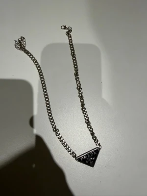 Prada black necklace | Unisex - Bilderna visar ett silvrigt halsband från Prada med en kedja och en triangel-formad berlock, men ingen väst finns med.