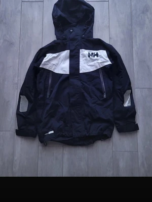 Svart och vit vindjacka Helly Hansen - Snygg svart och vit vindjacka från Helly Hansen med huva och dragkedjor framtill. Jackan är i storlek 152 men passar om man brukar ha storlek xs. Skriv för fler bilder💕