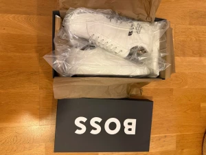 Boss skor i läder (Gary tenn) HELT NYA  - Stilrena vita sneakers från BOSS med klassisk låg profil och rund tå. Skorna är tillverkade i läder och har vita snören samt diskret branding på sidan. Perfekta för en clean och modern look. Helt nya i original förpackning. Ny pris 3599 mitt pris 1000kr först till kvarn!