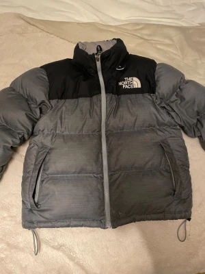 Grå och svart pufferjacka North Face - Säljer en grå och svart pufferjacka från The North Face med hög krage och två sidofickor med dragkedja. Jackan har en tydlig logga på bröstet och ryggen, samt justerbara snören vid nederkanten. Perfekt för kalla dagar och har en clean, urban vibe.