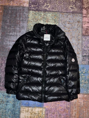 Svart Moncler pufferjacka - Svart pufferjacka från Moncler med glansig finish och klassisk logga på ärmen. Jackan har hög krage, dragkedja framtill och sidofickor med dragkedja. Insidan har en unik tvättinstruktionsetikett med serietidningsmotiv. Perfekt för dig som vill ha en snygg och varm jacka.