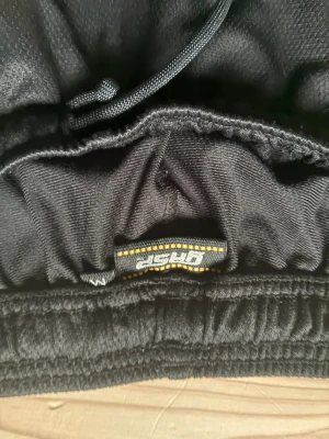 Svarta GASP mjukisbyxor med tryck - Säljer ett par svarta mjukisbyxor från GASP med resår och snörning i midjan. Byxorna har ett stort grått GASP-tryck och orange logga på ena benet. Perfekta för gymmet eller chill hemma. Materialet känns mjukt och bekvämt.