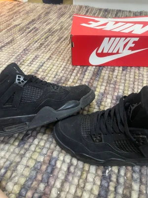 Nike Air Jordan 4 Retro Black Cat - Säljer ett par Nike Air Jordan 4 Retro Black Cat i helsvart mocka och mesh. Klassisk siluett med Jumpman-logga på hälen och tungspetsen. Skorna har svarta snören, plastdetaljer och en chunky sula för extra street vibe. Perfekta för dig som gillar stilrena och ikoniska sneakers. De kommer med extra snören. De är storlek 39 men funkar som 40!
