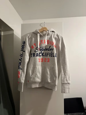 Grå Superdry hoodie - Säljer en grå hoodie från Superdry med stor tryckt text i blått och rött på bröstet och ärmen.