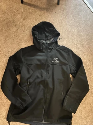 Svart vindjacka från Arcteryx  - Svart vindjacka från Arc'teryx med huva och dragkedja framtill. Jackan har två sidofickor med dragkedja och är tillverkad i vattenavvisande syntetmaterial med mjukt grått fleecefoder på insidan. Snygg logga på bröstet och justerbar huva för extra skydd mot väder.