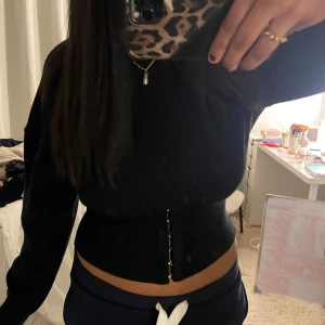 Svart croppad korsett-topp - Säljer en svart croppad korsett-topp med lång ärm och snygga hakknäppen framtill. Toppen har en tight passform och markerar midjan på ett stilrent sätt. Perfekt för dig som vill ha en edgy och trendig look.