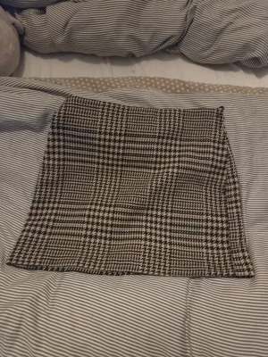 Rutig kortkjol med dragkedja - Snygg kortkjol med rutigt mönster i svart, beige och vitt. Kjolen har en rak passform och är gjord i ett tjockare material som känns som ull eller syntet. Stängs med en diskret dragkedja i sidan. Lite använd men i nästan nyskick