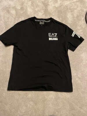 Svart EA7 Emporio Armani t-shirt - Svart t-shirt från EA7 Emporio Armani med coolt tryck fram och bak. Framsidan har logga och Milano-text, baksidan har SEVEN FEVER i stora bokstäver och schackrutigt band. Kortärmad modell i mjuk bomull med vita detaljer på ena ärmen.