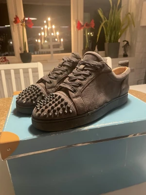 Christian Louboutin grå mocka sneakers - Säljer ett par grå sneakers från Christian Louboutin i mocka med svarta nitar på tån och klassisk röd sula. Skorna har rund tå, snörning och en platt sula. Insidan är fodrad med beige skinn för extra komfort. Perfekt för dig som gillar edgy och lyxig stil.(man får box o kvitto kan skicka bilder på mer)