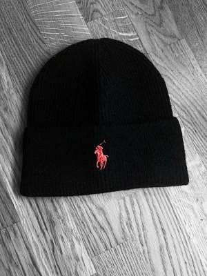 Svart stickad mössa från Polo Ralph Lauren - Svart ribbstickad mössa från Polo Ralph Lauren med klassisk röd broderad logga framtill. Mössan har uppvikt kant och är gjord i mjukt stickat material som håller dig varm och snygg under kalla dagar.