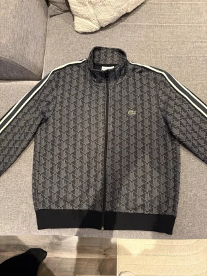 Svart mönstrad track jacket Lacoste - Snygg svart track jacket från Lacoste med geometriskt mönster och vita ränder längs ärmarna. Jackan har dragkedja framtill, ståkrage och klassisk krokodillogo på bröstet. Köpt på johnells för 2300 mitt pris 800. Hör av er vid funderingar.