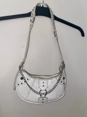 Vit axelväska med kedjedetaljer  - ” smuts färgen” är köpt så. Cool vit axelväska i skinnimitation med silvriga kedjor och nitar. Väskan har dekorativa sömmar, dragkedja upptill och justerbart axelband med metallringar. Perfekt för dig som gillar edgy och trendiga accessoarer.