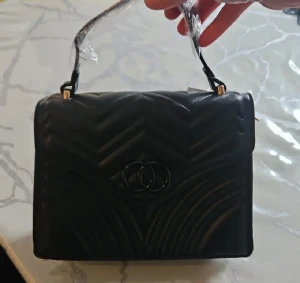 Svart quiltad handväska med mönster - Stilren svart handväska i quiltad design med snyggt mönster och dekorativt spänne framtill. Väskan har ett kort handtag med gulddetaljer och är tillverkad i skinnimitation. Perfekt för dig som vill ha en trendig accessoar med attityd.