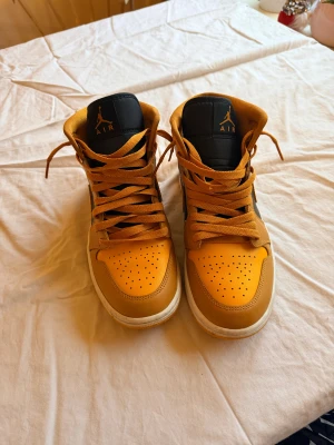 Nike Air Jordan 1 Mid gul/svart - Säljer ett par Nike Air Jordan 1 Mid i en riktigt snygg orange och svart färgkombination. Skorna har klassisk snörning, perforerad tåbox och vit mellansula. Tillverkade i skinn med ikoniska Jordan Wings-loggan på sidan och Jumpman på plösen. Perfekta för dig som vill sticka ut med dina sneakers. Obs. Absolut nyskick - burits max 3 ggr.
