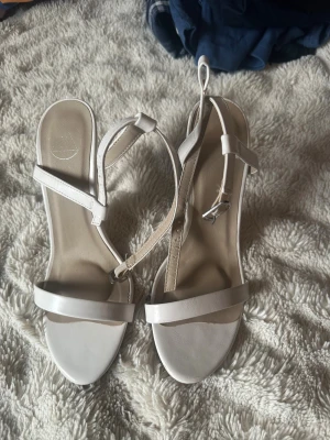 Vita sandaletter med hög klack - Säljer ett par stilrena vita sandaletter med hög klack och smala remmar. Skorna har öppen tå och justerbart spänne runt ankeln. Tillverkade i syntetmaterial med en elegant och minimalistisk look som passar perfekt till sommarens outfits.