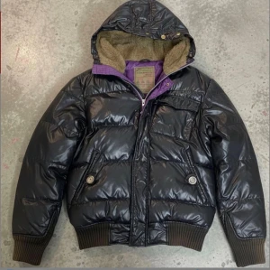 Japansk Archive pufferjacka från PPFM - Säljer en svart pufferjacka från PPFM med glansig yta, lila innerfoder och mjuk beige fuskpäls i huvan. Jackan har ribbade muddar, dragkedja, knappar och flera fickor. Det kommer också med päls för luvan