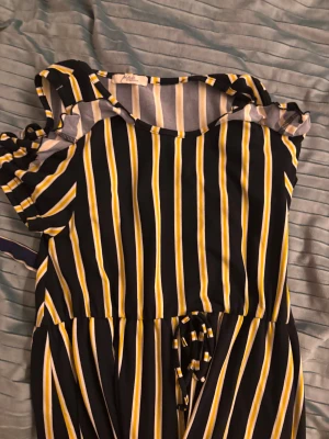 Randig kortklänning från pino premium - Superfin kortklänning med svarta, vita och gula ränder från Vero Moda. Klänningen har korta ärmar med volangdetalj och knyt i midjan för en snygg passform. Perfekt för dig som gillar färg och mönster!