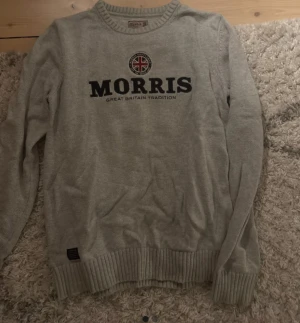 Grå stickad tröja från Morris - Snygg grå stickad tröja från Morris med rund hals och ribbade muddar. Stor logotyp med brittisk flagga på bröstet och texten 'Great Britain Tradition'. Perfekt för dig som gillar klassisk stil med en modern touch. Använd väldigt fåtal gånger.