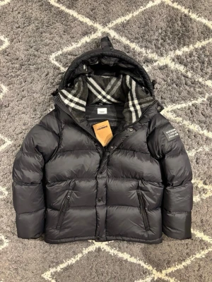 Svart Burberry pufferjacka  - Svart pufferjacka från Burberry med avtagbara ärmar, så du kan bära den som väst. Jackan har huva med snörning, dragkedja och knappar framtill samt två fickor med dragkedja. Insidan har klassiskt Burberry-rutigt foder i grått och vitt. Perfekt för kalla dagar. Liten i storlek.