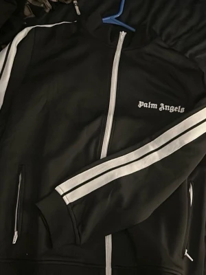 Palm Angels kofta - Svart track jacket från Palm Angels med vit dragkedja och vita ränder längs ärmarna. Jackan har hög krage och logga tryckt på bröstet. Tillverkad i mjuk polyester för en sportig och clean look.