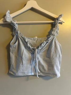 Ljusblå linnetopp med spets från NLY Trend - Supersöt ljusblå linnetopp från NLY Trend i storlek M. Toppen har breda axelband med spetsdetaljer och en snygg snörning framtill som ger en cool v-ringad look. Materialet är mjukt och känns skönt mot huden, perfekt för varma dagar.
