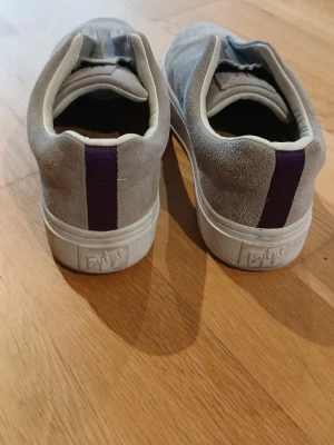 Grå mockasneakers från Eytys - Snygga grå sneakers från Eytys i mocka med vit sula och en lila detalj baktill. Skorna har en minimalistisk design utan snörning och en korkinnersula med Eytys-logga. Perfekta för dig som gillar stilrena och bekväma skor. Pris går att diskuteras.