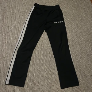 Palm Angels strl M kom med pris förslag  - Svarta mjukisbyxor från Palm Angels med vita sidoränder och logotyp på vänster lår. Byxorna har elastisk midja. Byxorna är äkta 