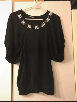 Svart blus med pärldetaljer storlek S/M - Säljer en svart blus med breda, korta ärmar och snygga pärldetaljer runt halsringningen. Blusen har en loose passform och är perfekt för dig som gillar stilrena plagg med lite extra bling. Passar till många olika looks!