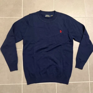 Marinblå tröja från Polo Ralph Lauren - Snygg marinblå tröja från Polo Ralph Lauren med röd broderad logga på bröstet och RLX-broderi på ärmen. Rund halsringning och ribbade muddar vid ärmslut och nederkant. Perfekt för dig som gillar klassisk stil med en sportig touch.