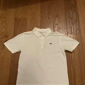 Vit pikétröja från Lacoste Sport - Vit pikétröja från Lacoste Sport med klassisk krage och korta ärmar. Tröjan har den ikoniska gröna krokodilen broderad på bröstet och knäppning framtill. Tillverkad i bomull för en skön och sportig känsla. Perfekt för dig som gillar stilren och enkel design. Storlek medium.