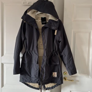 Svart parkajacka från Tretorn - Parkajacka från Tretorn i svart med beige teddyfoder på insidan. Jackan har huva, snörning i midjan, stora fickor med lock och dragkedja framtill. Materialet är polyester och jackan har en längre bakdel för extra skydd mot väder. Perfekt för kalla dagar.