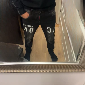 Svarta Dsquared2 ICON jeans med tryck - Säljer ett par svarta Dsquared2 ICON jeans med vit text på framsidan och coola färgstänk-detaljer. Byxorna har klassisk femficksdesign, kedjedetalj och smal passform. Perfekta för dig som vill sticka ut med en edgy streetstyle.