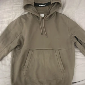 Grön hoodie från C.P. Company - Säljer en grön hoodie från C.P. Company med klassisk linsdetalj på ärmen. Tröjan har huva med snörning, magficka och ribbade muddar. Perfekt för dig som gillar streetwear och vill ha en clean look med snygga detaljer. Storlek S, säljer för jag vuxit ur den. 9/10 skick. Ny pris 2500kr.