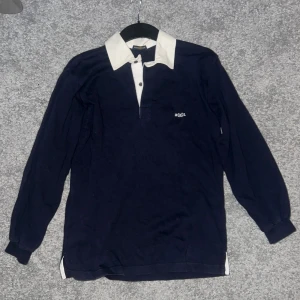 Marinblå pikétröja Henri Lloyd - Snygg marinblå pikétröja från Henri Lloyd med vit krage och knappslå. Tröjan har lång ärm och diskret broderad logga på bröstet. Tillverkad i 100% bomull för skön känsla och klassisk look. Perfekt för dig som gillar preppy stil.