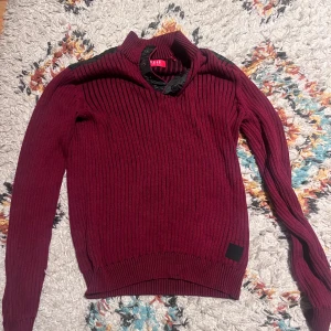 Vinröd ribbad tröja från Guess XS - Snygg vinröd ribbstickad tröja från Guess i storlek XS. Tröjan har svarta detaljer på axlarna och vid halsen, samt knappar vid kragen. Långärmad och med en cool, lite sportig look som sticker ut. Bra för grisig stil med tröjor inte många har . Storlek xs men passar storlek s också använd ett få par gånger