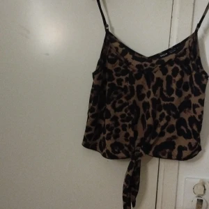 Leopardmönstrad linne-topp från SHEIN - Säljer en snygg leopardmönstrad linne-topp från SHEIN i storlek M. Toppen har tunna justerbara axelband, v-ringning och en knyt-detalj framtill. Färgerna är brun och svart, och materialet känns mjukt och lätt. Perfekt för dig som vill sticka ut med ett djurmönster!