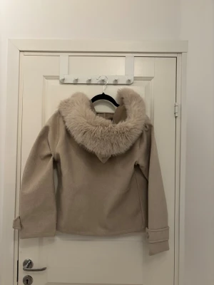 Beige kappa med fuskpäls och knappar - Säljer en beige kappa med fluffig fuskpäls runt kragen och huvan. Jackan har stora knappar framtill, två fickor och snygga detaljer vid ärmsluten. Perfekt för dig som vill ha en stilren och mysig look under höst och vinter.