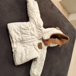 Vit teddyjacka med huva från SHEIN - Söt vit teddyjacka för baby från SHEIN med mjuk brun teddyfoder i huvan och gulligt nallemärke på bröstet. Jackan har dragkedja framtill, ribbade muddar och quiltad design. Perfekt för kyliga dagar och håller ditt barn varmt och mysigt.