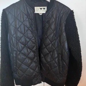 Svart quiltad bomberjacka Junkyard - Svart bomberjacka från Junkyard med quiltad framsida i syntet och teddyärmar. Jackan har dragkedja framtill, ribbade muddar och en cool kontrast mellan glansigt material och fluffiga ärmar. Perfekt för dig som gillar streetstyle och vill sticka ut.