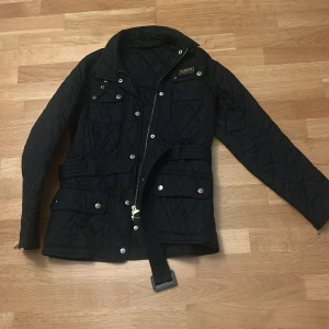 Svart quiltad jacka från BARBOUR - Quiltad svart jacka från Barbour med bälte i midjan, flera fickor med tryckknappar och dragkedja framtill. Klassisk design med Barbour-logga på bröstet och snygga detaljer i silver. Perfekt för dig som gillar stilren och funktionell look.