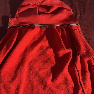 Röd fleecejacka från Sail Racing - Riktigt schysst röd fleecejacka från Sail Racing med huva och dragkedja. Jackan har mjukt fleecefoder på insidan och ribbade muddar. Perfekt för dig som gillar sportig stil och vill ha något som värmer under kyliga dagar.