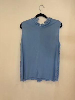 Blå ärmlös blus med spetsdetaljer - Supersöt ljusblå ärmlös blus med blommig spets på ryggen och runt halsen. Framsidan är slät medan baksidan har ett vackert spetsmönster som ger en romantisk vibe. Perfekt för dig som gillar detaljer och vill sticka ut lite extra.