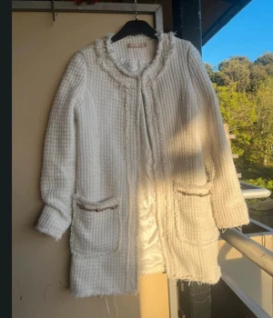 Vit kappa med fickor och fransdetaljer - Stilren vit kappa i grovstickat material med fransade kanter och två stora fickor framtill, dekorerade med kedjedetaljer. Långärmad och rak passform, perfekt för dig som gillar en klassisk men trendig look.