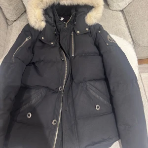 Svart Moose Knuckles dunjacka med päls - Säljer en svart dunjacka från Moose Knuckles med fluffig päls på huvan, coola metalldetaljer och dragkedjor. Jackan har flera fickor med knappdetaljer och snyggt broderat märke på insidan. Perfekt för dig som vill ha en varm och stilren look i vinter. Ny pris 13000kr köpt på jb 