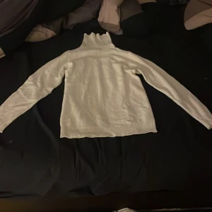 Vit polotröja med ribbad detalj - Säljer en vit polotröja med lång ärm och hög krage. Tröjan har en snygg ribbad detalj som går längs mitten både fram och bak, samt ribbade muddar vid nederkant och hals. Perfekt för dig som gillar stilrena och klassiska plagg.