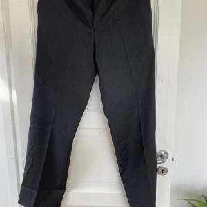 Mörkgrå kostymbyxor från Oscar Jacobson - Snygga mörkgrå kostymbyxor från Oscar Jacobson i 100% ull. Klassisk rak passform med pressveck och diskreta bakfickor med knappar. Perfekt för dig som gillar stilrena och tidlösa plagg med exklusiv känsla.